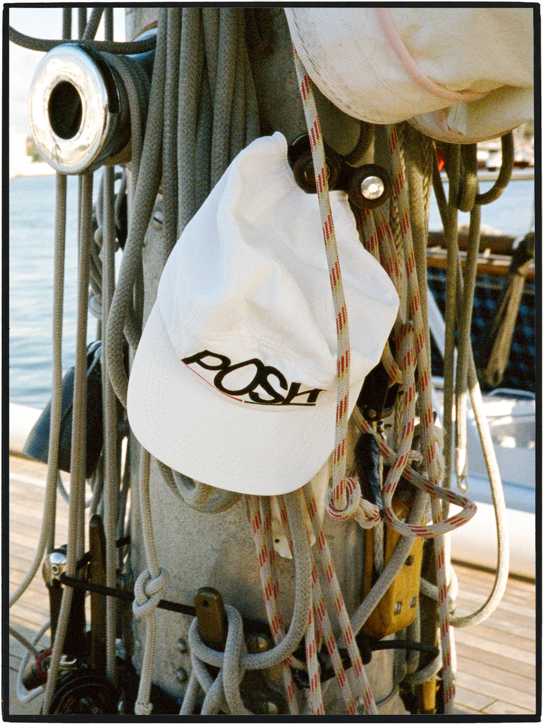 A white baseball cap with the word 'POSH' printed in black across the front, hanging among ropes and rigging on a boat deck.