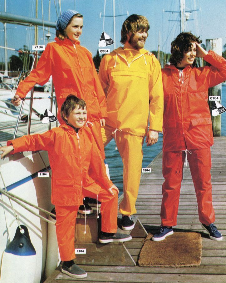 Four people wearing bright orange and yellow raincoats on a dock with boats in the background.