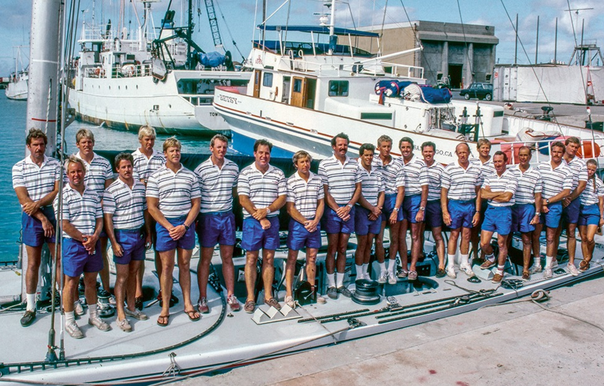 Group of people in striped shirts and blue shorts standing on a boat with ships in the background.