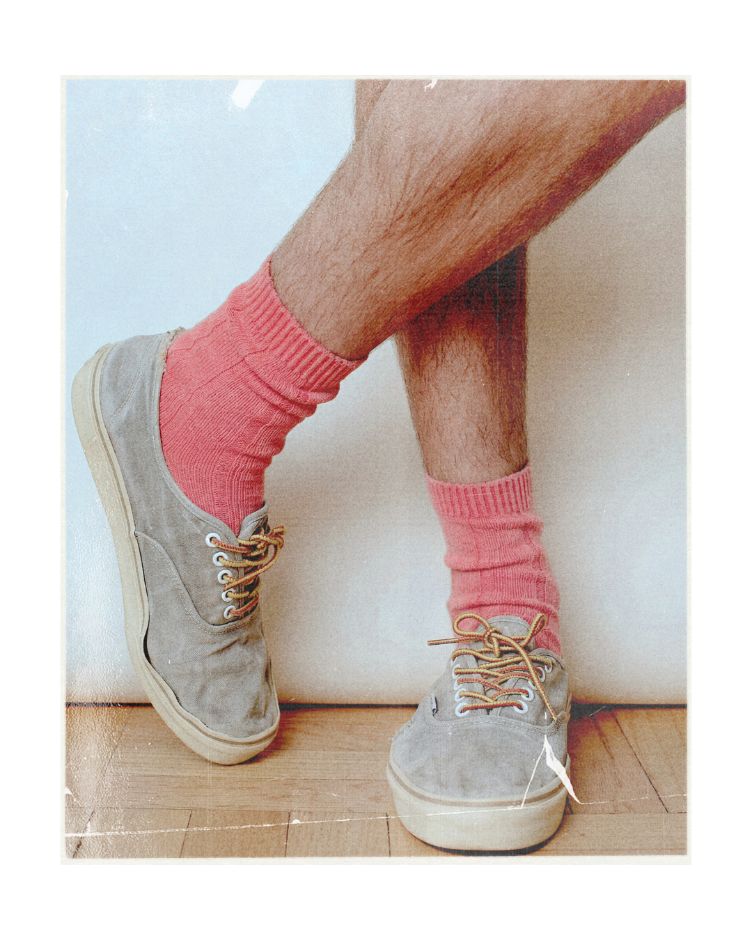 A person's legs crossed, wearing pink ribbed crew socks and light gray suede sneakers with tan laces, set against a light blue wall and wooden floor.