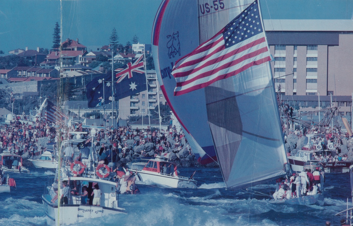 Sailboat with American flag on a body of water with spectators in the background