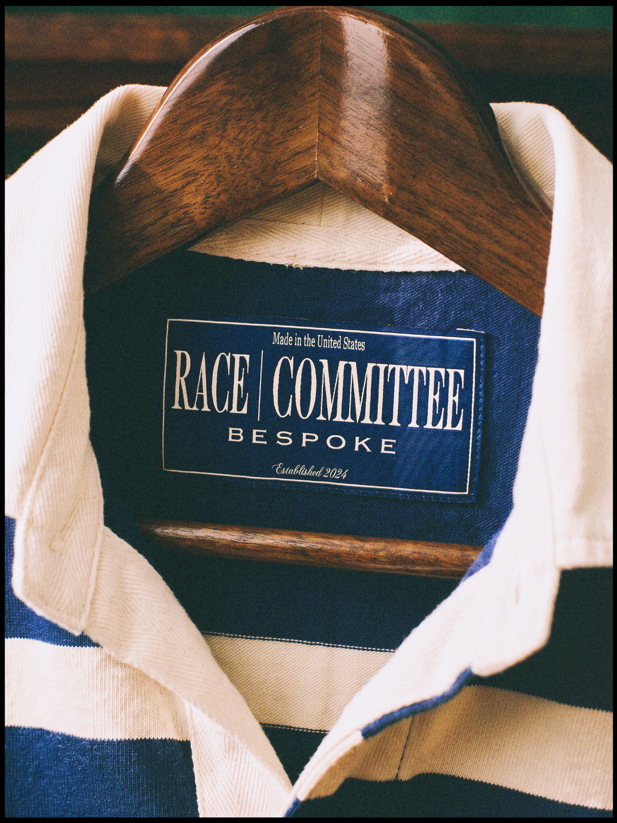A close-up of a navy and cream striped knit polo shirt collar on a wooden hanger, showing the blue and white 'RACE COMMITTEE BESPOKE' label.