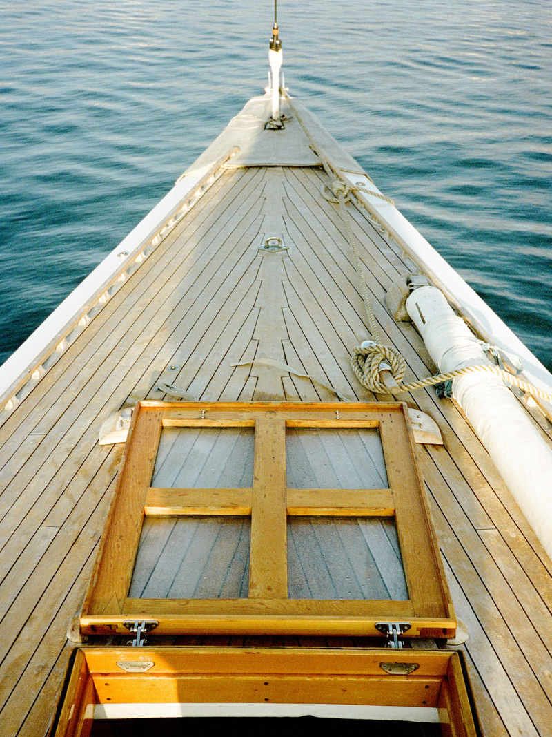 A close-up view from the deck of a wooden boat, looking forward toward the bow with blue water on both sides. A wooden hatch cover with glass panes is open in the foreground, and ropes and white rigging are visible along the side.