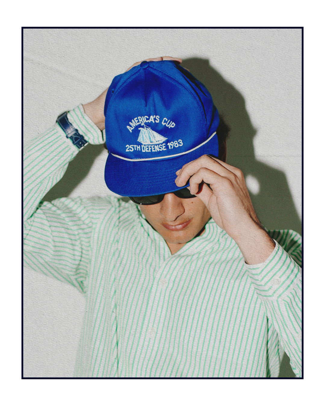 Person wearing a blue cap with text, green striped shirt, and sunglasses on a light background