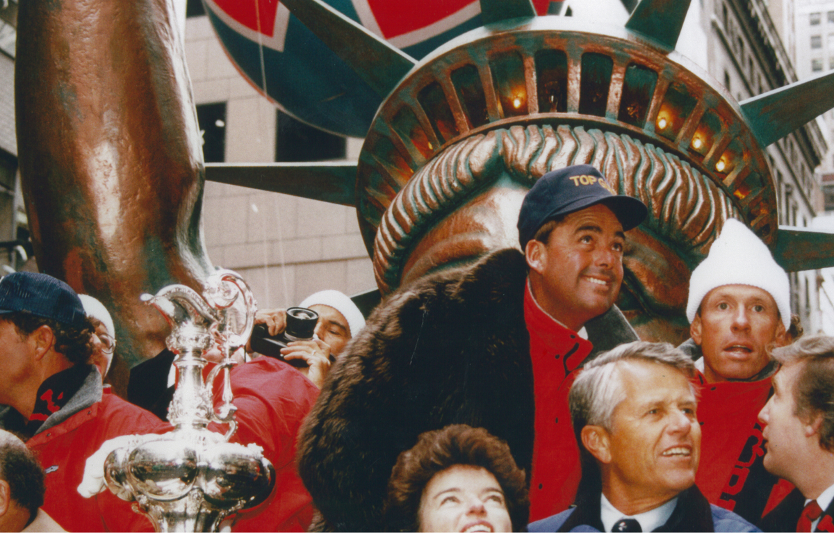 Group of people with a trophy in front of a large Statue of Liberty decoration, likely at a parade or event.