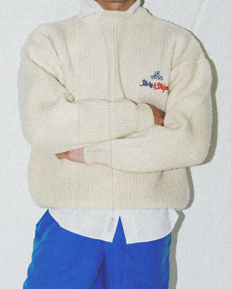 Person wearing a cream sweater with a logo, white shirt, and blue pants on a light background